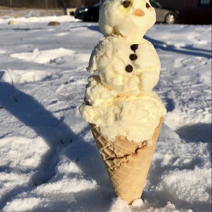 A snowman created using vanilla ice cream in a waffle cone that is sitting in the snow