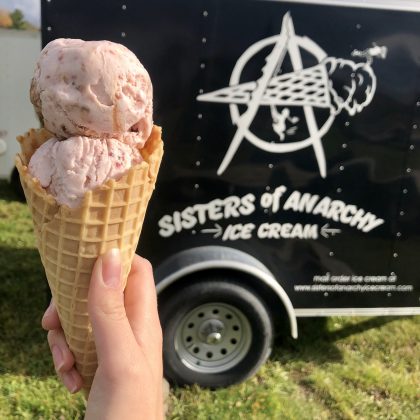 Two scoops of pink-speckled ice cream inside of a cone, held in front of a black trailer. White letters say Sisters of Anarchy Ice Cream beneath the company's logo