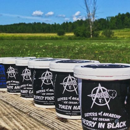 six black and white pints of ice cream sit on top of a wooden surface, with a background of green grass, trees and blue sky